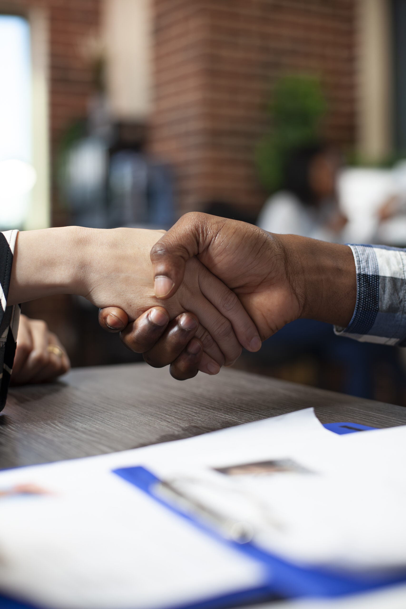 Closeup of multiethnic individuals handshaking over the office desk during job interview meeting. Selective focus on hands of diverse people doing a greeting gesture in brick wall workspace.